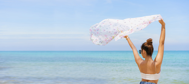 Beautiful Asian young woman in bikini enjoy walking and looking at the sea and beach in morning, woman rising a white scarf over her head.