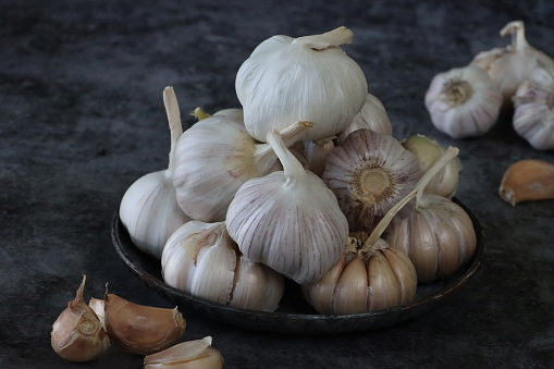 Stock photo showing close-up view of a stack of fresh, whole garlic bulbs on a mottled black plate, against a mottled black background.