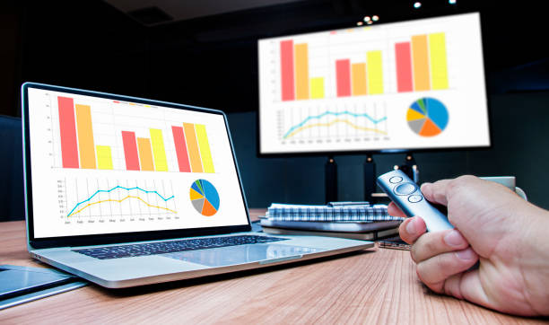 istock Businessman hand hold remote control slide with mock up chart slide show presentation on display laptop and television in meeting room 1442675349