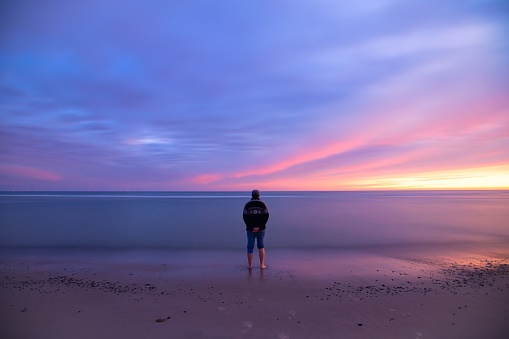 A back shot of an adult man stands at the beach looking at the sea and dusk sky