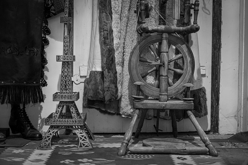 The grayscale view of a wooden spinning wheel, statuette, coats, and woman boots on the carpet of a room