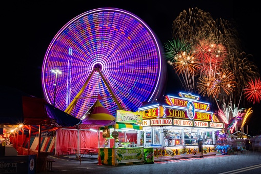 Harrington, United States – February 07, 2019: A colorful Ferris wheel and food stall lights against breathtaking fireworks during a Delaware State Fair