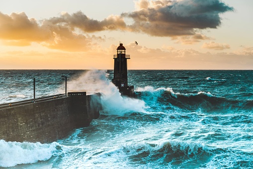 A large waves crash against the stone tower of the lighthouse at high tide at sunset