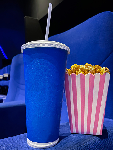 Stock photo showing close-up view of striped carton of popcorn in cinema / movie theatre. Caramelised / caramel toffee popcorn as movie snack food besides disposable cup and drinking straw in cinema with empty blue seats.