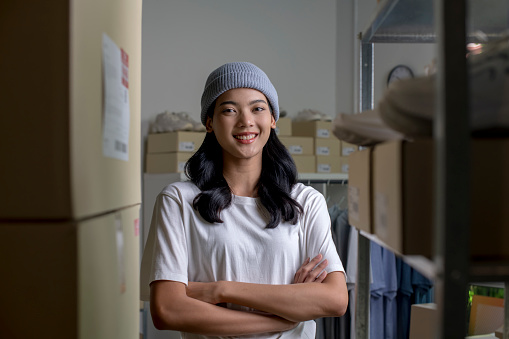 Confident young Asian woman retail seller, entrepreneur, online store drop shipping small business owner looking at camera standing in delivery shipping warehouse with parcel boxes.