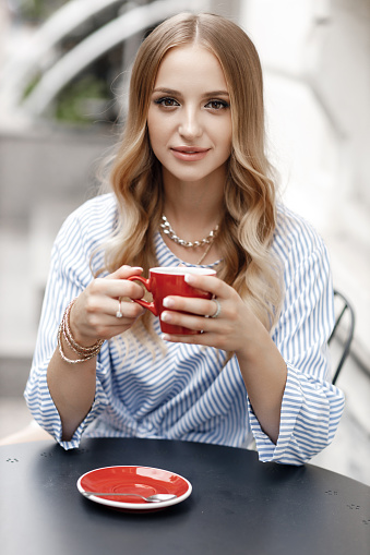 Portrait of a beautiful girl with blond curly hair outdoors, sitting at a round table in a street cafe, holding a cup of coffee. Pretty smiling young woman in a good mood is sitting at a table in an outdoor cafe.Summer holiday, vacation concept.