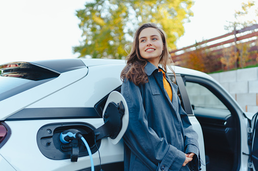 Young woman waiting while electric car charging in home charging station, sustainable and economic transportation concept.