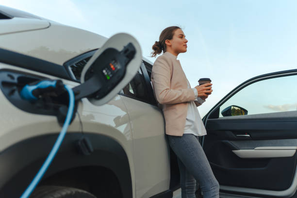 Jeune femme avec une tasse de café attendant pendant que sa voiture électrique recharge, concept de transport durable et économique. - Photo