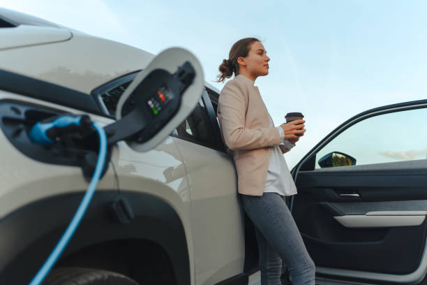 Young woman with cup of coffee waiting while her electric car charging, sustainable and economic transportation concept. stock photo