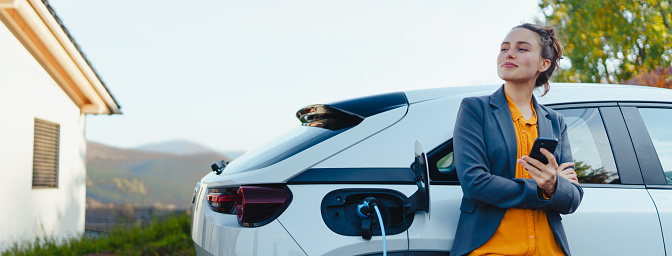 Young woman with smartphone waiting while electric car charging in home charging station, sustainable and economic transportation concept.