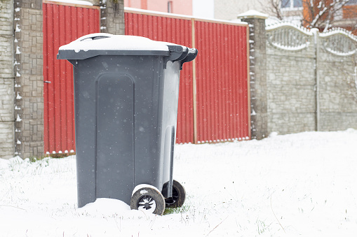 A trash can covered with snow next to a house. Garbage removal in winter