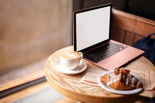 Freelance workplace in cafe. Table on coffeeshop with open laptop, newspaper and morning breakfast, notebook computer with cup of coffee and croissant