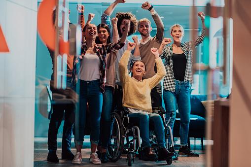 Photo of business women in wheelchairs with their hands raised in the air with their colleagues, together celebrating business success.