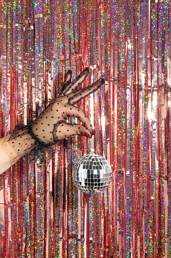 Christmas and New Year creative layout with woman hand in black lace glove holding disco ball decoration infont of foil fringe pink curtains background. 80s or 90s retro fashion aesthetic celebration or party concept.