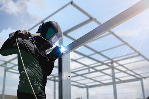 Male worker using steel pole welding machine, blue sparks from welding, wearing mask and gloves for construction safety. blurred steel structure background