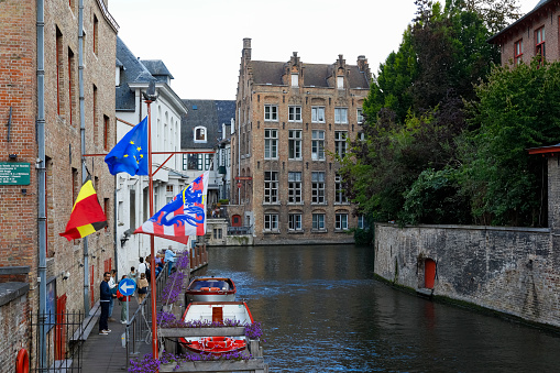 Bruges, Belgium - September 8, 2022: There are brick buildings by the canal, next to them there is a small pier on which there are people, two boats are moored by the pier