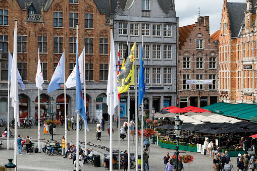Bruges, Belgium - September 15, 2022: Large traditional brick houses surround the Market Square with outdoor restaurants and cafes, and flagpoles with flags. Usually, there are a lot of people here