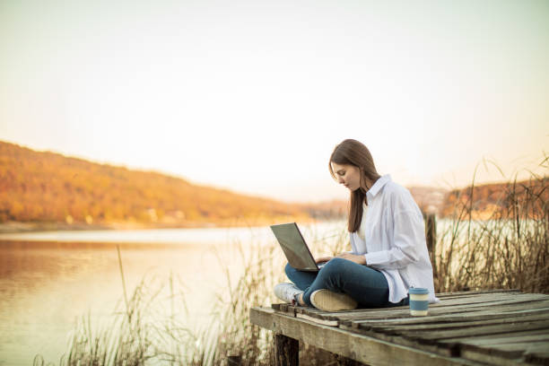 istock Young beautiful woman using laptop outside by lake 1442730168