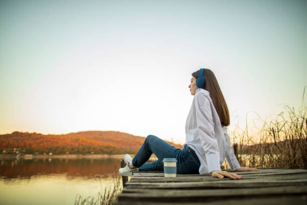 Young woman resting on wooden pier by lake. She is listening music drinking coffee and enjoying nature. Beautiful woman is wearing jeans and white shirt. Lake water is colored with orange from sunset at autumn.