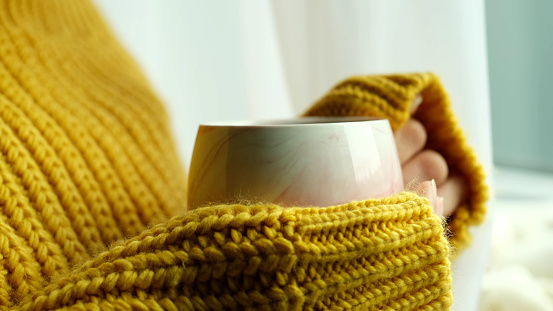 unrecognizable woman in a warm knitted sweater holds a cup of hot coffee in her hands. atmosphere of coziness and comfort in your home.