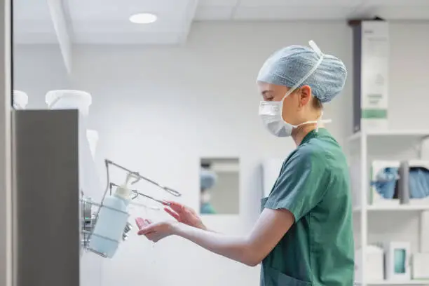 Side view of a woman nurse wearing surgical mask and cap using hand sanitizer at hospital. Antiseptic liquid for hand washing in operation room.