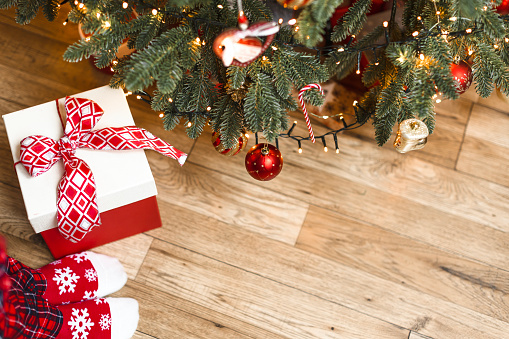 A girl in red socks stands near a Christmas tree with a gift. Christmas background with decorated fir and space for a copy.