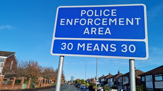 Blue UK road traffic sign with a white boarder and white letters reading police enforcement area 30 means 30 standing along side a road in a built up residential area reminding drivers not to speed.