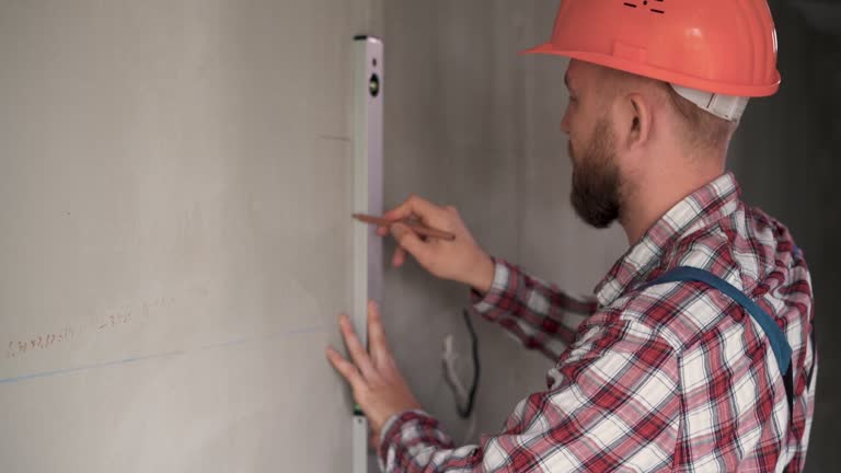 male engineer or architect in a protective helmet measures the level of the wall. A builder in a house is checking a flat concrete wall. Repair and construction