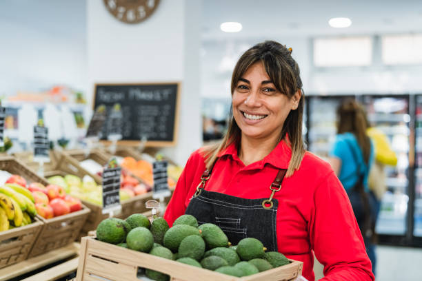 Latin woman working in supermarket holding a box containing fresh avocados stock photo