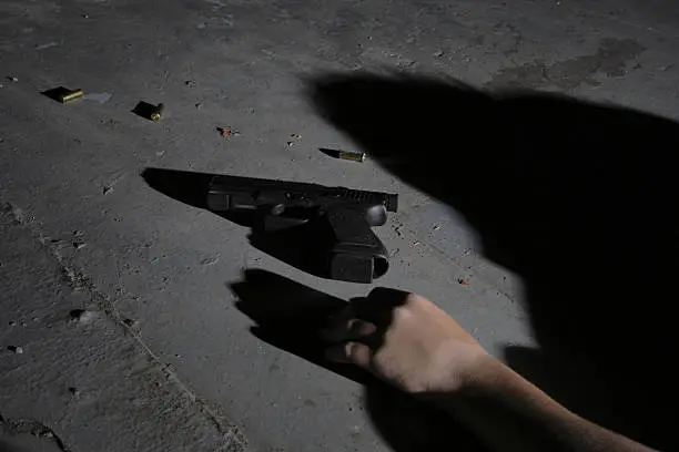 A semi-automatic hand gun lying on a concrete warehouse floor with spent shells and a limp hand next to it, implying a shooting.