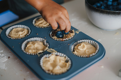 Close-up view of child's hands decorating the still raw muffins with blueberries. Funny lovely healthy child having fun with helping. Helping parents