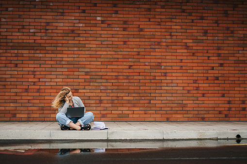 Front view of a student girl with curly hair sitting down on a street near the university working on a calculator while posing against the background of a red brick wall with free space for text. Copy space. People lifestyle. Happy lifestyle.