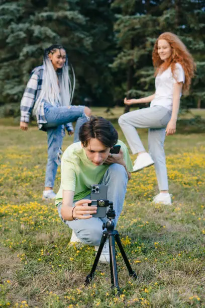 Photo of Teenagers dancing at camera filming video using cell phone on tripod at city park, creating trendy content. Gen Z Entertainment. Contemporary art.