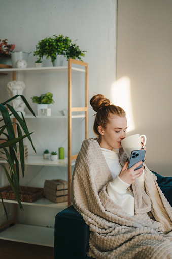 Caucasian woman wrapped in warm cozy plaid holding mug of tea, enjoying leisure at home while chatting with someone on mobile phone. Domestic lifestyle. Entertainment and leisure.