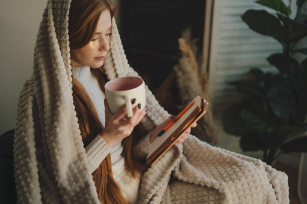 istock Young red-headed woman drinking tea and reading a book while resting on sofa in the weekends. Wrapped in a blanket. Beauty treatment. Body care. Beauty routine. 1442780088