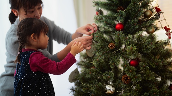 A mother and her preschool age daughter are decorating a Christmas tree in the living room at home.