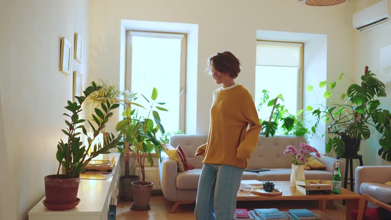 Kobieta ubrana w żółty sweter i niebieskie dżinsy stoi w przytulnym, pełnym roślin salonie.
