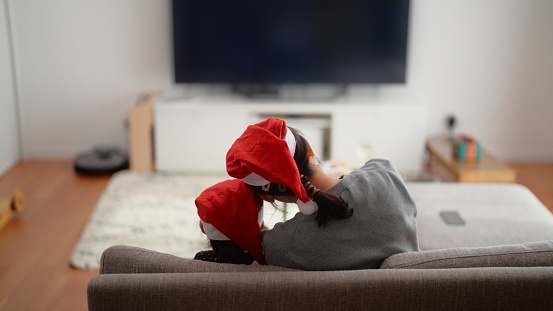 A mother and her small preschool age daughter are enjoying watching tv together in the living room at home during Christmas.