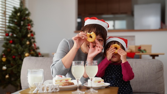 Portrait of mother and her small daughter enjoying Christmas and making funny faces and looking through doughnuts hole at camera at home
