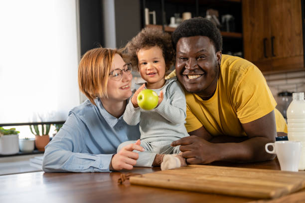 multicultural family with a cute child at home kitchen. parents feeding a small daughter with fruit healthy food - domestic life young family family child imagens e fotografias de stock