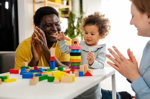 Mother and father supporting their cute little daughter in playing with colorful didactic wooden toys at home