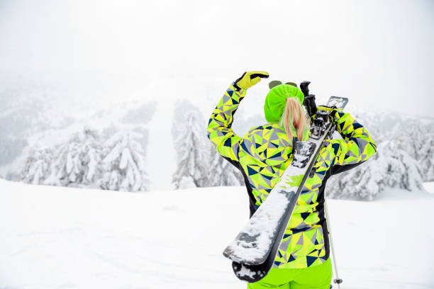 Back of Woman skier in cold winter day with ski equipment in mountain hill. Female in lime color reflective sports wear on winter Holiday vacation. Copy space