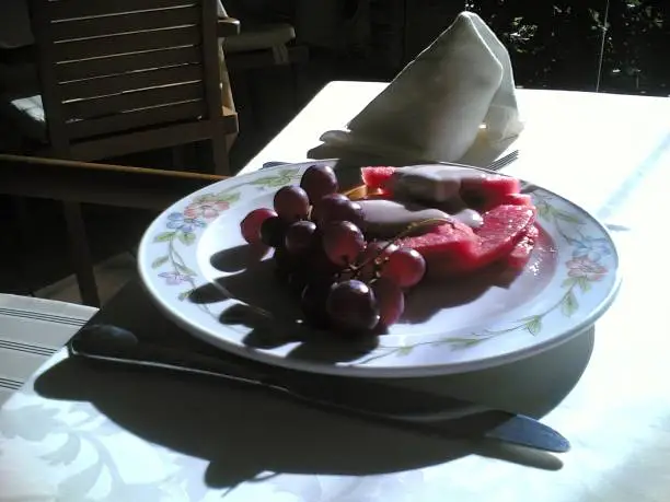 Healthy fruit breakfast on the terrace of a Santo Domingo hotel.
A branch of grapes and slices of watermelon with cream in the frame, on a plate next to cutlery.
In the background is a figuratively twisted napkin.
Santo Domingo, Dominican Republic.