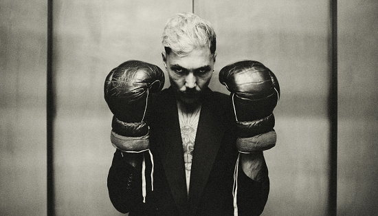 Portrait of a young man with boxing gloves. He scowls at the viewer, wears a jacket and has a tattoo on his chest. He is in front of a closed elevator door.