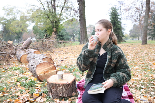 Beautiful young girl reads a book and drinks coffee sitting on a blanket in the park