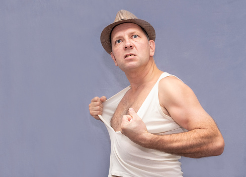 Middle-aged Caucasian man in a hat. Strong emotions, angry and tearing his shirt.