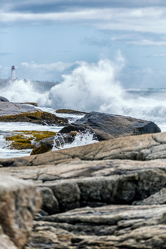Distant Peggy's Cove lighthouse stands tall far beyond the Atlantic ocean coastline storm surf crashing onto and between the granite rock formations and boulders at water's edge in the foreground. Nova Scotia, Maritime Provinces, Canada.