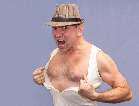 Middle-aged Caucasian man in a hat. Strong emotions, angry and tearing his shirt.