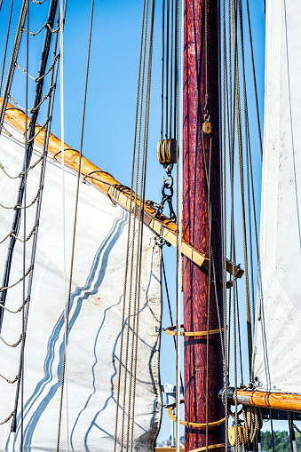 Close-up detail of intricate rigging lines, sails, pulleys and hooks, rope ladders, sail booms and masts as a large sail is being hoisted into place on an old tall ship sailing schooner being readied to cast off and sail at sea.