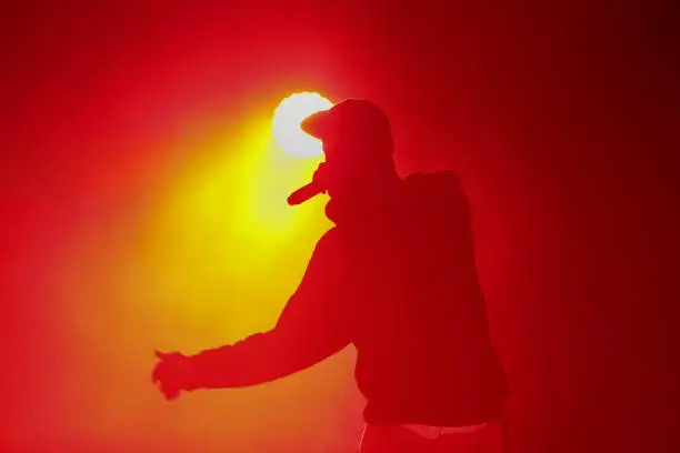 Photo of Silhouette of rap singer with microphone. Cool rapper with mic in hand singing on concert stage in bright red lights. Hip hop artist performing live on scene in music hall